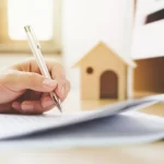 picture of a hand writing on a piece of paper with a wooden model of a house in the background