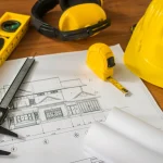 Image of blueprints, hard hat, leveler, measuring tape and construction headphones.